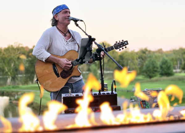 Picture taken from low angle in front of flames of firepit of male country blues cover singer for parties performing at outdoor event | Altus Entertainment